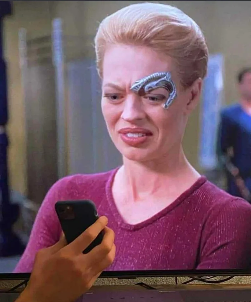 A woman with a metallic, ridged eyebrow implant looks down at a smartphone with a disappointed expression. She wears a…