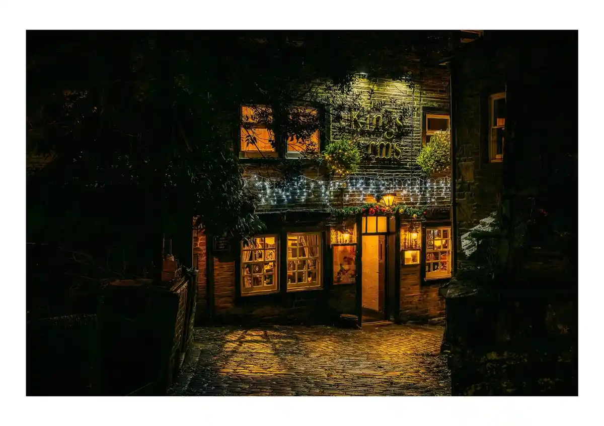 A nighttime photo of a cozy, warmly lit stone pub named 'The Kings Arms,' decorated with string lights and festive garlands…
