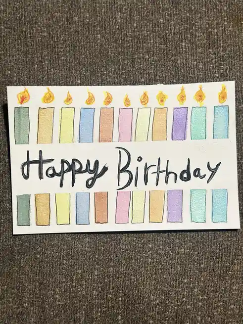 A hand-drawn birthday card on white paper shows ten colorful candles with flames at the top and identical candles without…