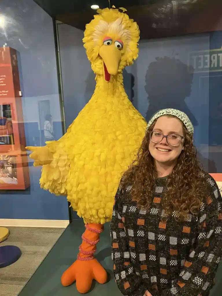 A woman with curly hair, glasses, and a patterned sweater smiles beside a large yellow Big Bird puppet on display. The…
