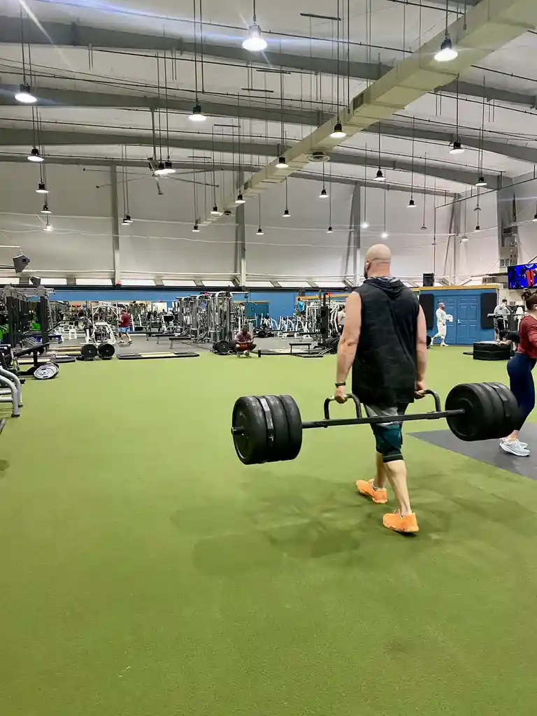 A bald man in a black sleeveless hoodie and bright orange shoes walks away from the camera through a large gym, carrying a…