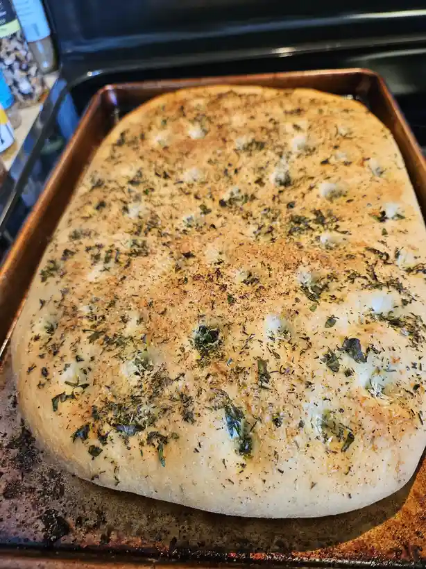 A close-up photo shows a rectangular, golden-brown baked flatbread on a well-used metal baking sheet, topped with herbs and…