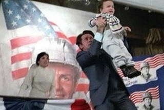 A man in a suit holds a baby high in the air while standing before a large American flag and a banner featuring a portrait…