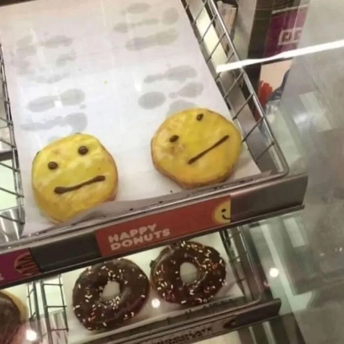 Two donuts in a bakery display case are decorated with icing to resemble faces: one with a smiling expression and one with a…