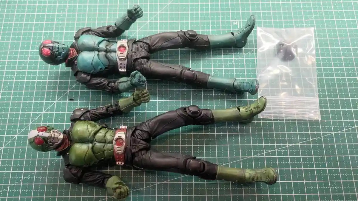 Two Kamen Rider Kuuga action figures lie on a green grid mat, one with teal chest armor and the other with green chest…
