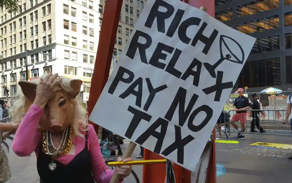 A person wearing a pig mask and pink top holds a protest sign reading 'RICH RELAX PAY NO TAX' with a martini glass icon…