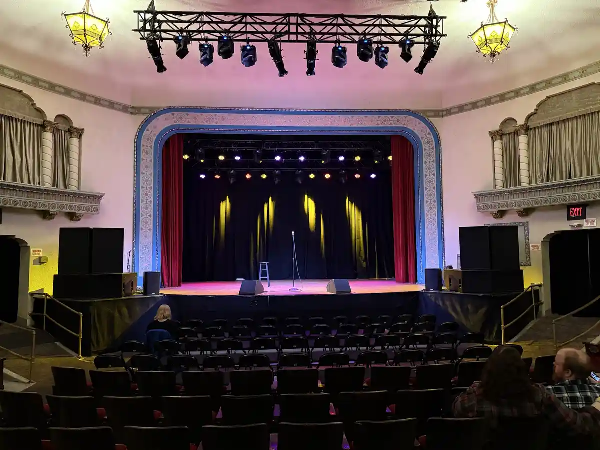 An empty theater stage with red curtains, spotlights, and a single microphone stand, viewed from the audience seating. A few…
