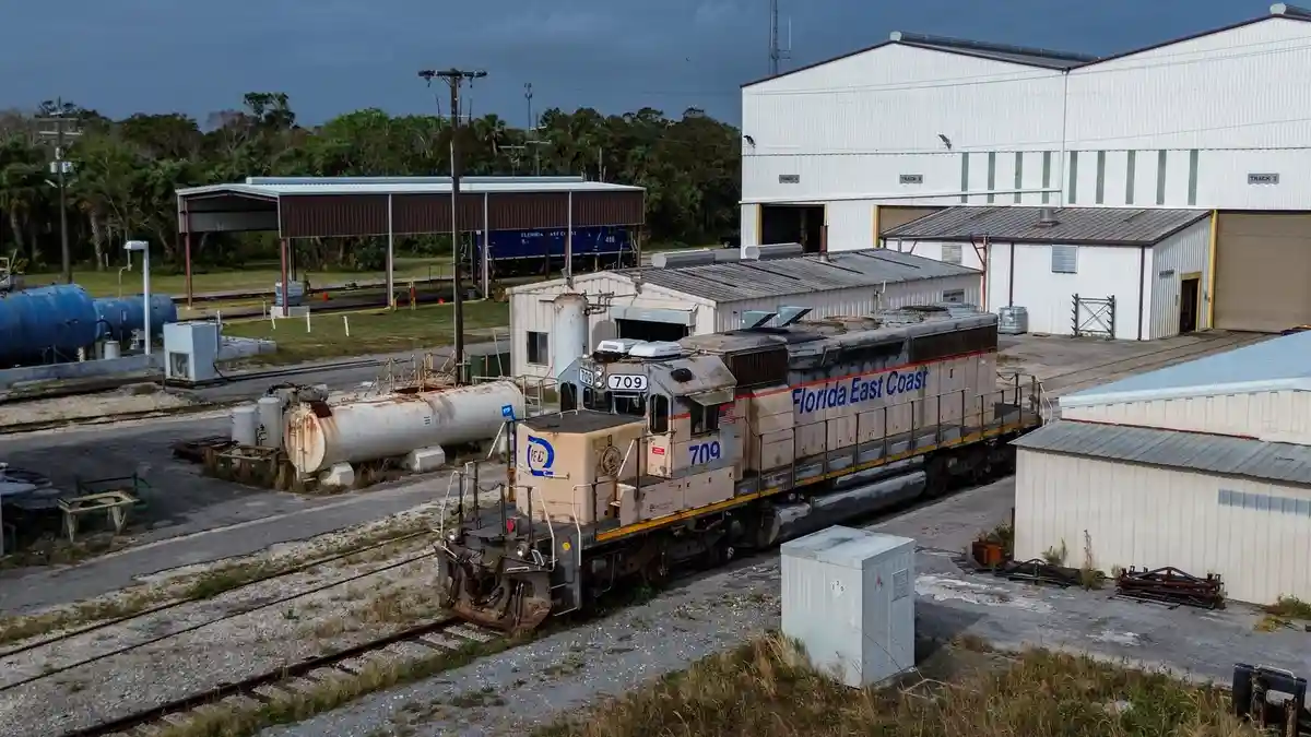 A weathered Florida East Coast locomotive, number 709, sits idle on tracks beside industrial buildings and rusted tanks…