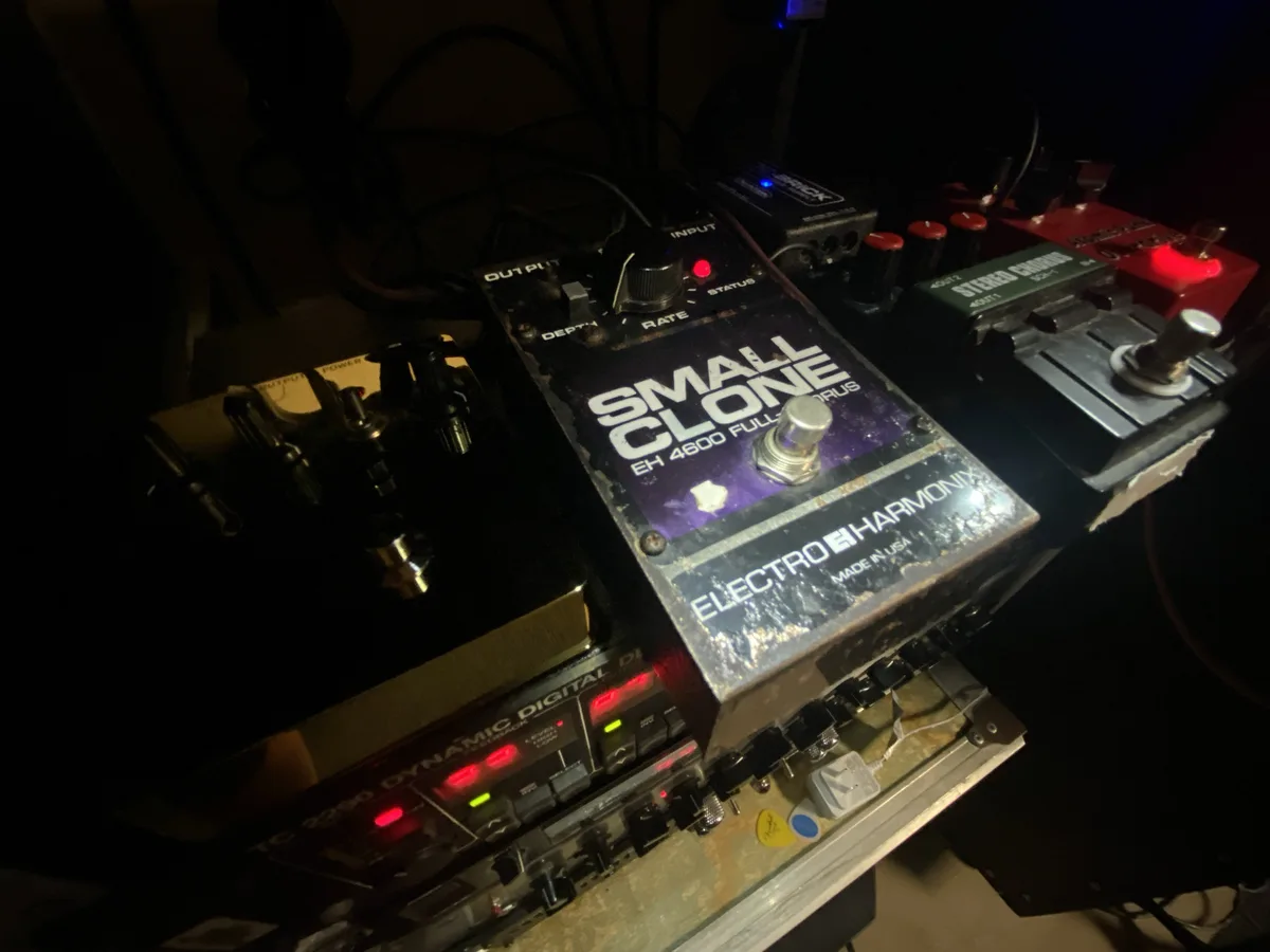 A close-up photo of a worn, dusty Electro-Harmonix 'Small Clone' guitar effects pedal, part of a cluttered pedalboard with…