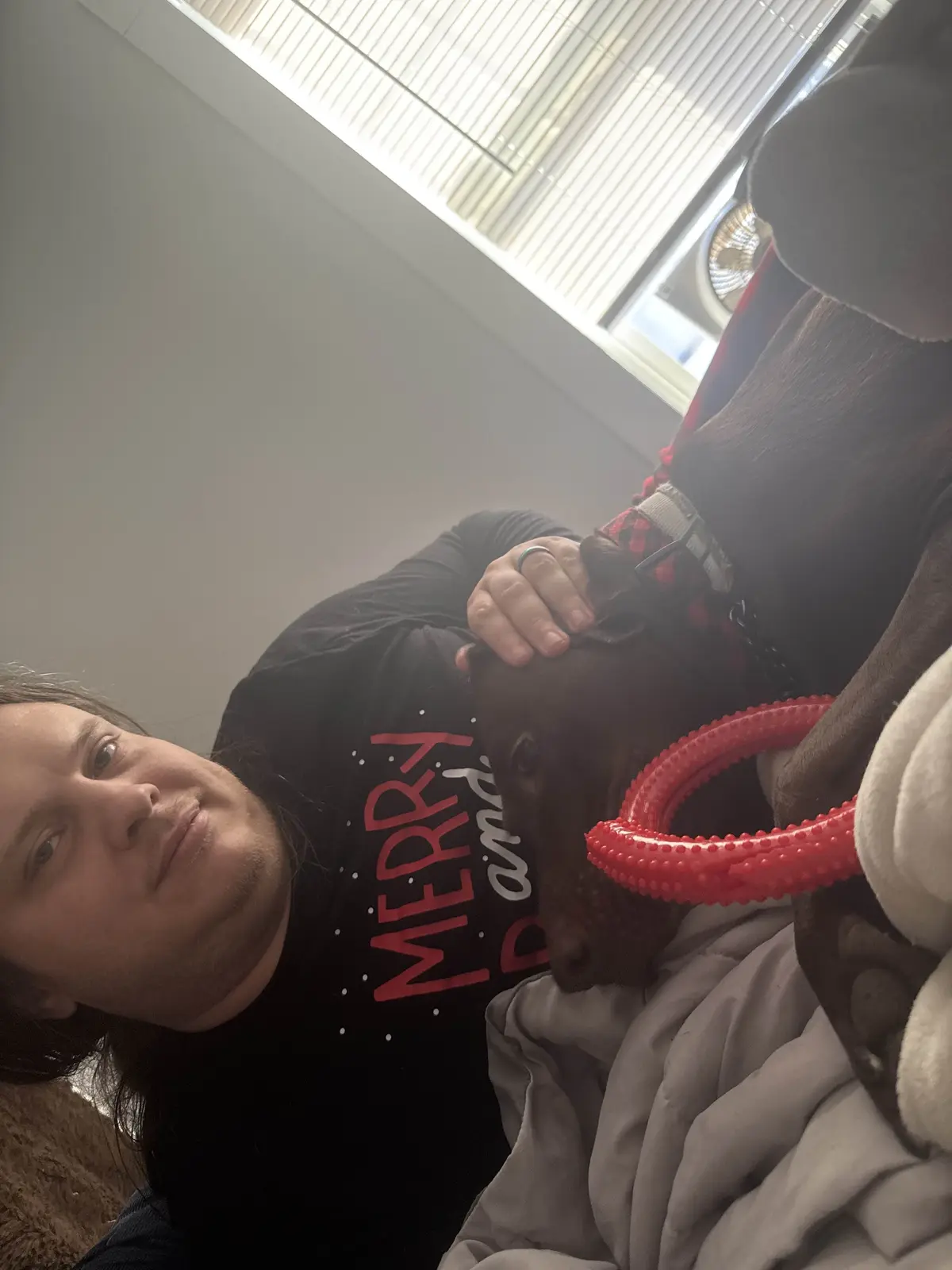 A person with long hair wearing a black t-shirt that says 'MERRY and bright' in red and white text is lying down and petting…