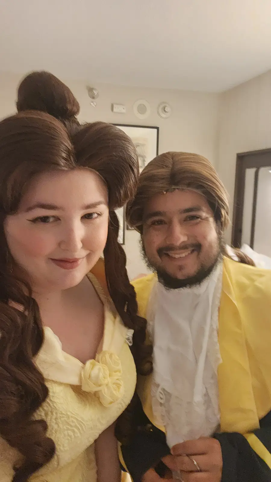 Two people dressed as Belle and the Beast from Beauty and the Beast pose for a selfie indoors. The woman wears Belle's…