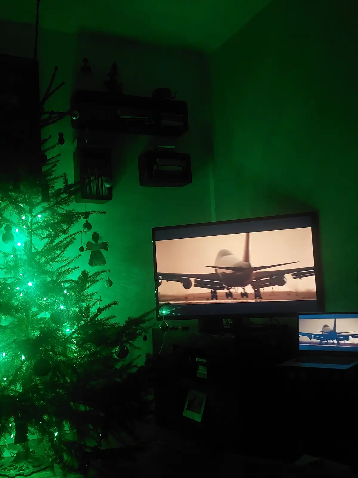 A dimly lit room bathed in green light, featuring a decorated Christmas tree with glowing lights on the left and two screens…