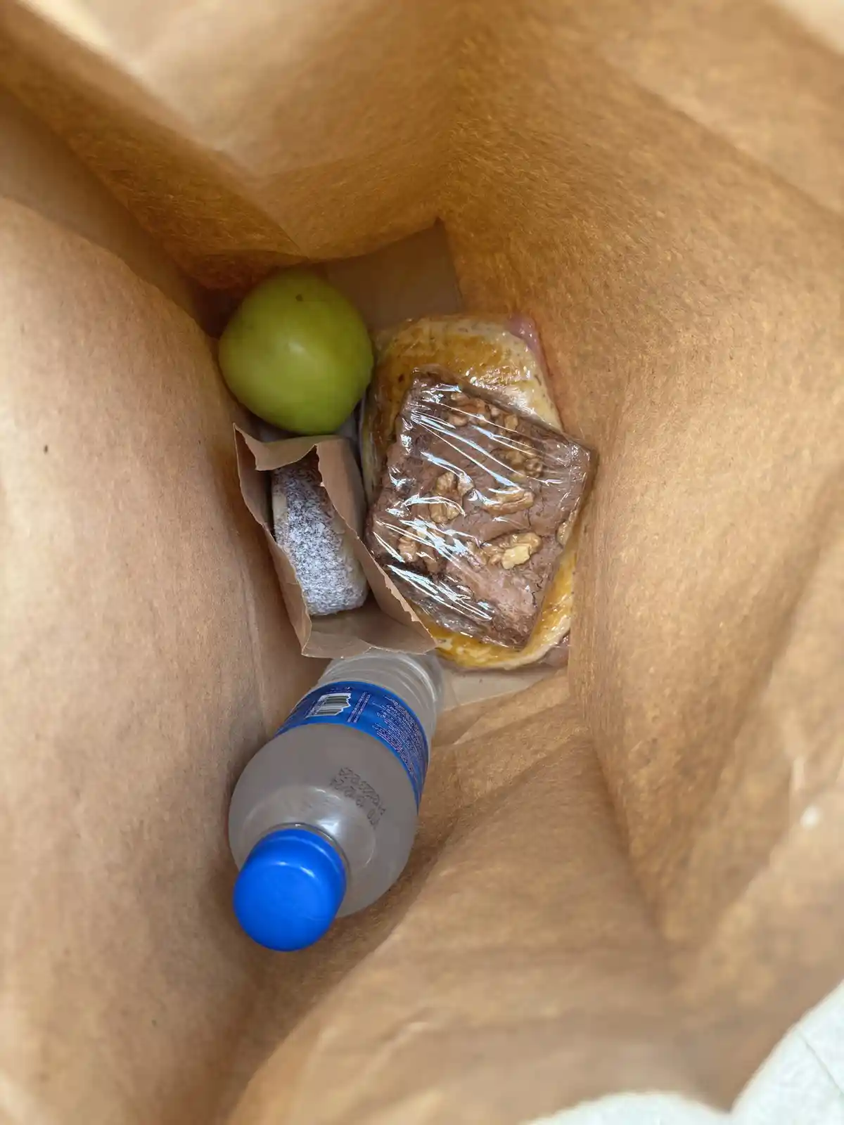 A top-down view of a brown paper bag containing a green apple, a wrapped chocolate brownie with walnuts, a sandwich, a…