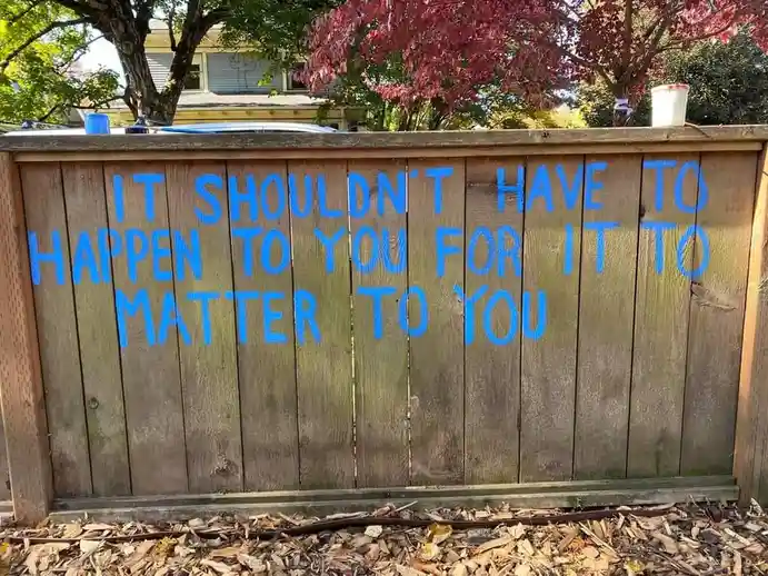 A wooden fence in a yard has blue spray-painted text reading 'IT SHOULDN'T HAVE TO HAPPEN TO YOU FOR IT TO MATTER TO YOU'…