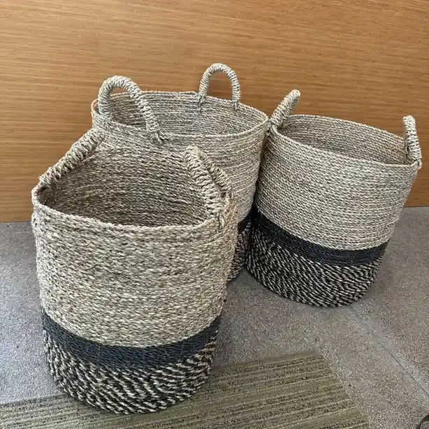 Three woven seagrass baskets with black and beige stripes sit on a tiled floor against a wooden wall. Each basket has two…
