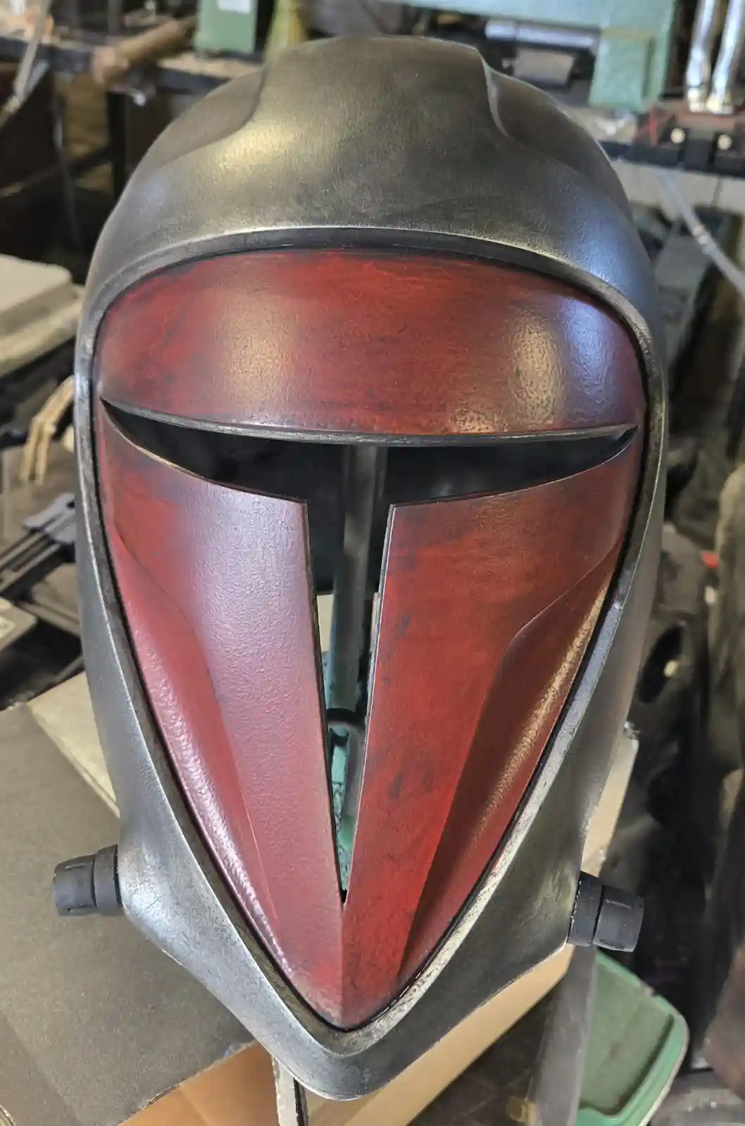 A close-up photo shows a custom-made Mandalorian helmet, featuring a dark grey metallic exterior and a red-tinted visor with…