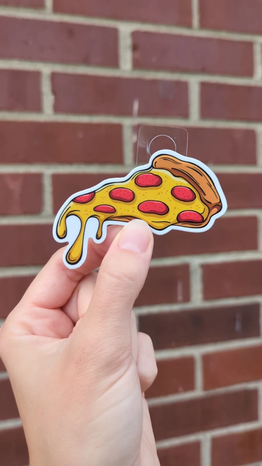 A hand holds up a cartoon sticker shaped like a slice of pepperoni pizza with melted cheese dripping down, against a red…