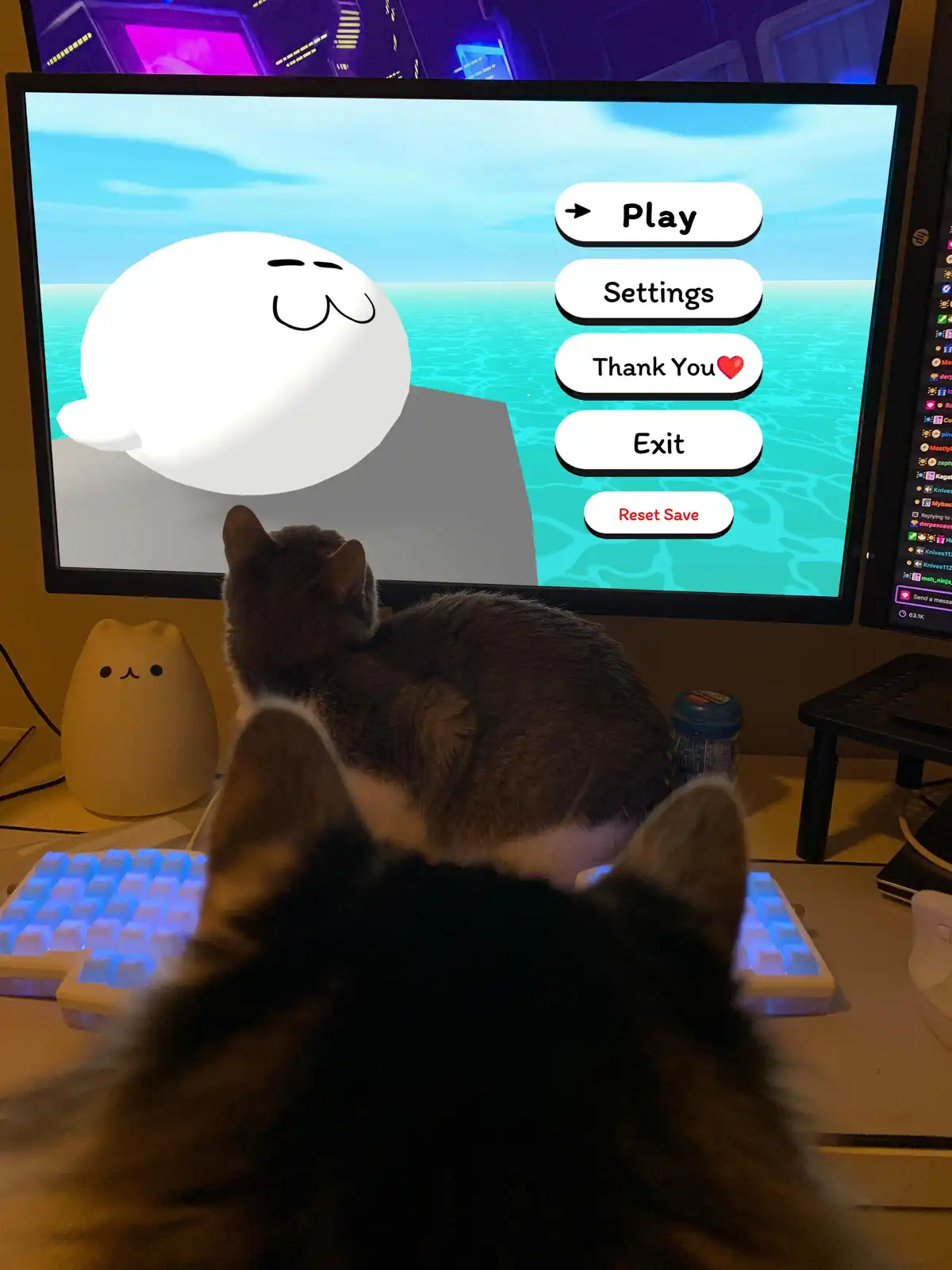 A real cat and a blurry husky-like dog are seen from behind, staring intently at a computer monitor displaying the main menu…