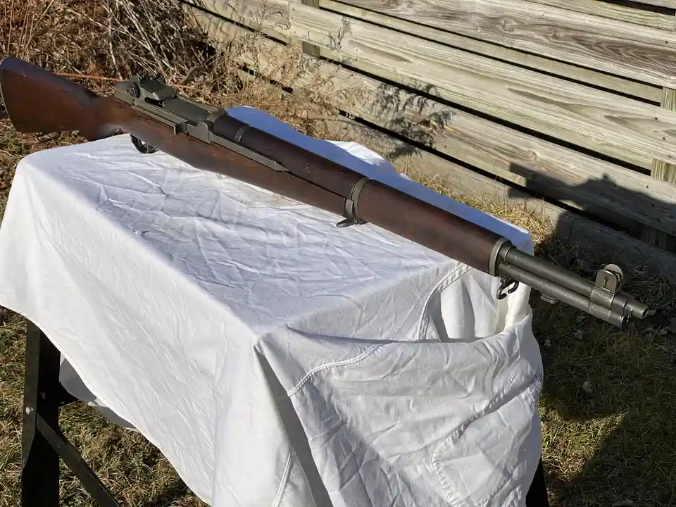 A vintage-style bolt-action rifle with a wooden stock and metal barrel rests diagonally on a white cloth-covered table…
