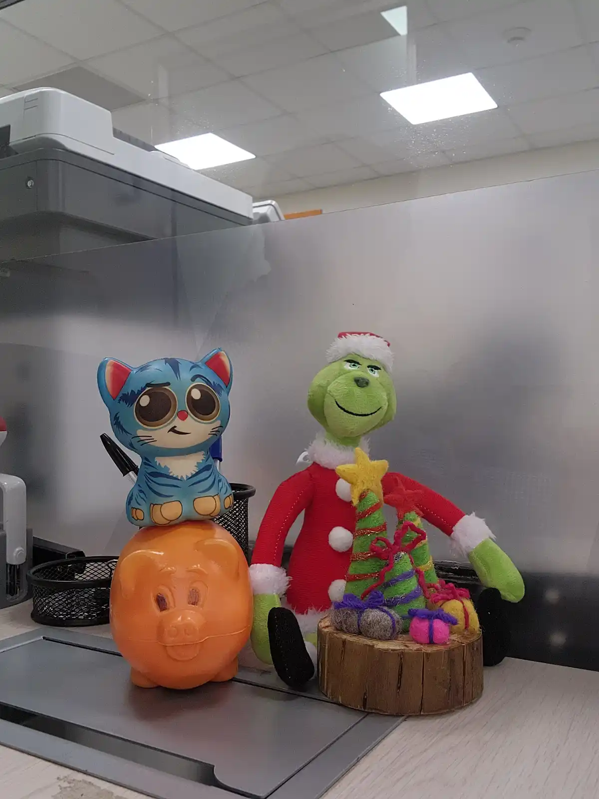 An office desk features a plush Grinch in a Santa suit holding a small felt Christmas tree, next to a blue cartoon cat…