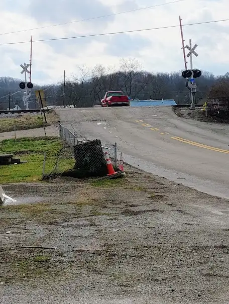 A red car is driving up a steep, gravelly hill toward a railroad crossing with active warning lights and gates. The road is…