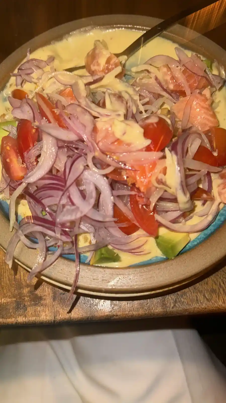 A close-up photo of a bowl of food containing salmon, cherry tomatoes, sliced red onions, avocado chunks, and a creamy…