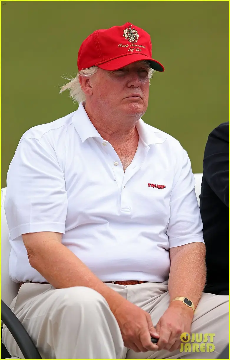 A man with blond hair wearing a red 'Trump International Golf Club' cap and a white polo shirt with 'TRUMP' embroidered on…