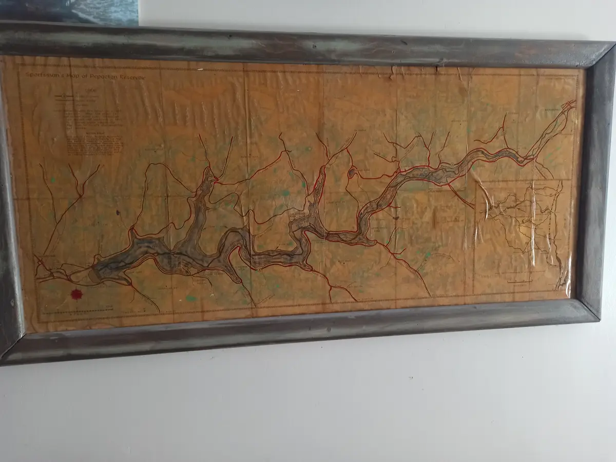 A framed, aged, sepia-toned map titled 'Sportsman's Map of Pepacton Reservoir' hangs on a white wall. The map shows winding…