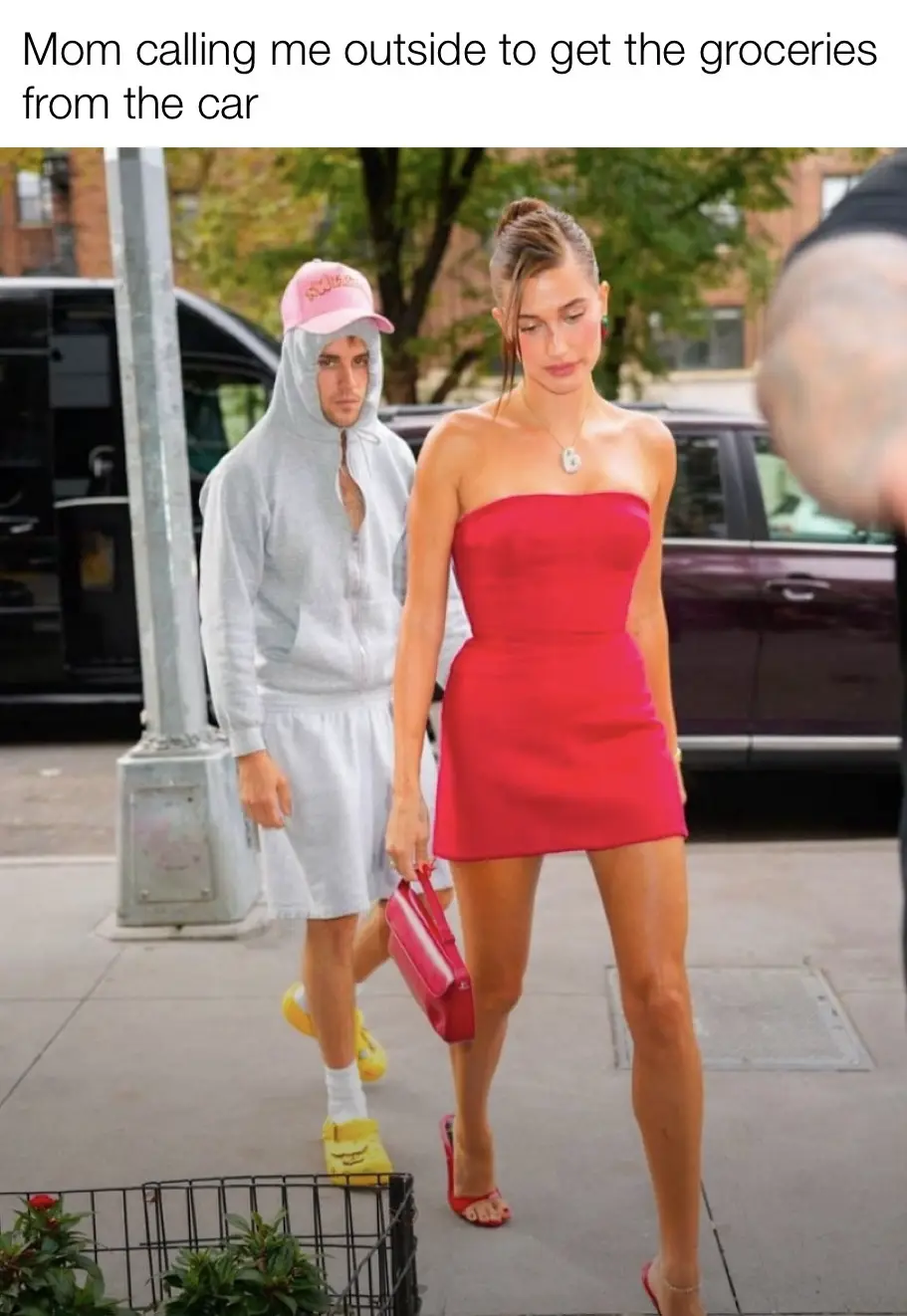 A man in a grey hoodie and pink cap walks beside a woman in a red dress, both on a sidewalk near parked cars. The overlaid…