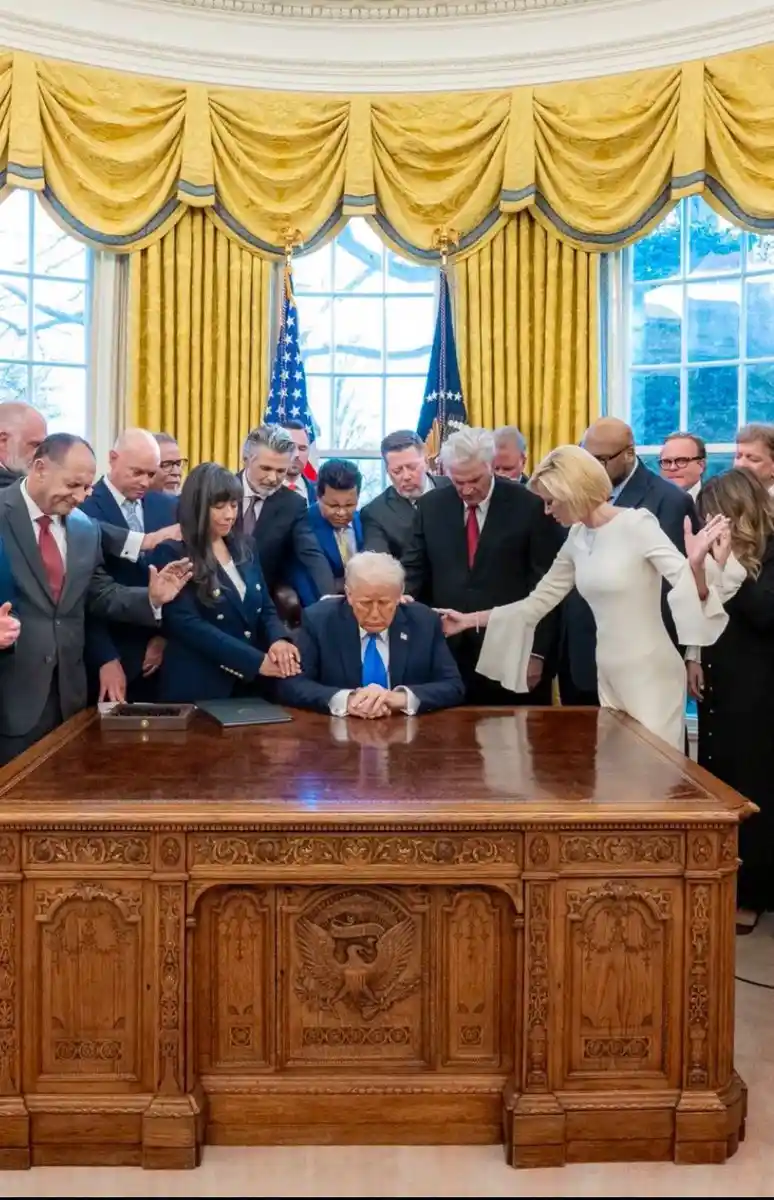 A man in a suit sits at a large wooden desk in an ornate room with yellow drapes and flags, surrounded by a crowd of people…
