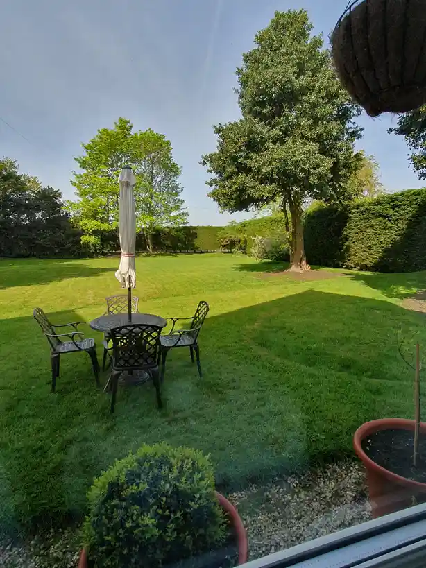 A view from a window shows a well-kept backyard with a round metal patio table and four matching chairs, a closed white…