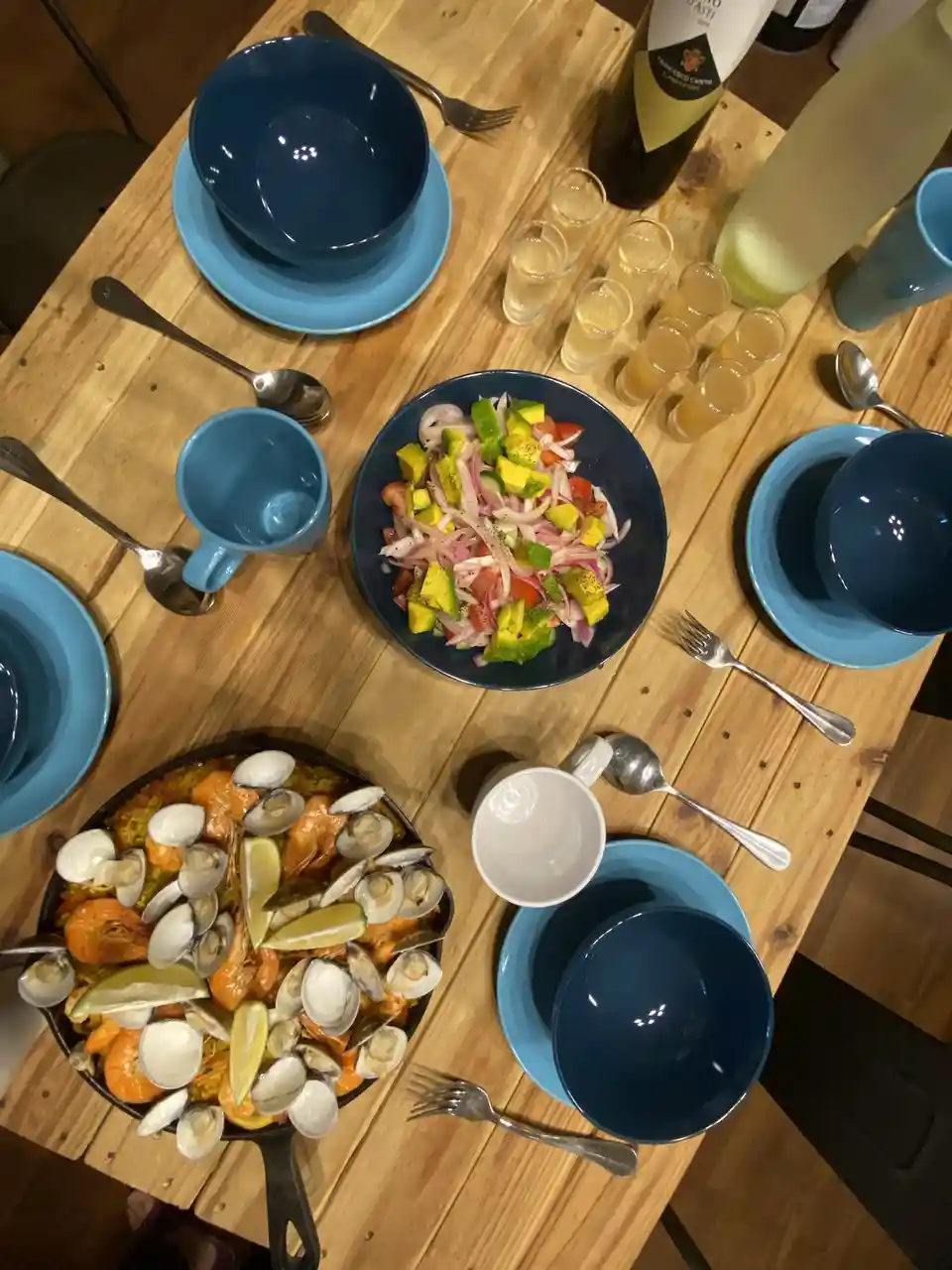 An overhead shot of a wooden table set for a meal, featuring a large skillet of seafood paella with clams and shrimp, a bowl…