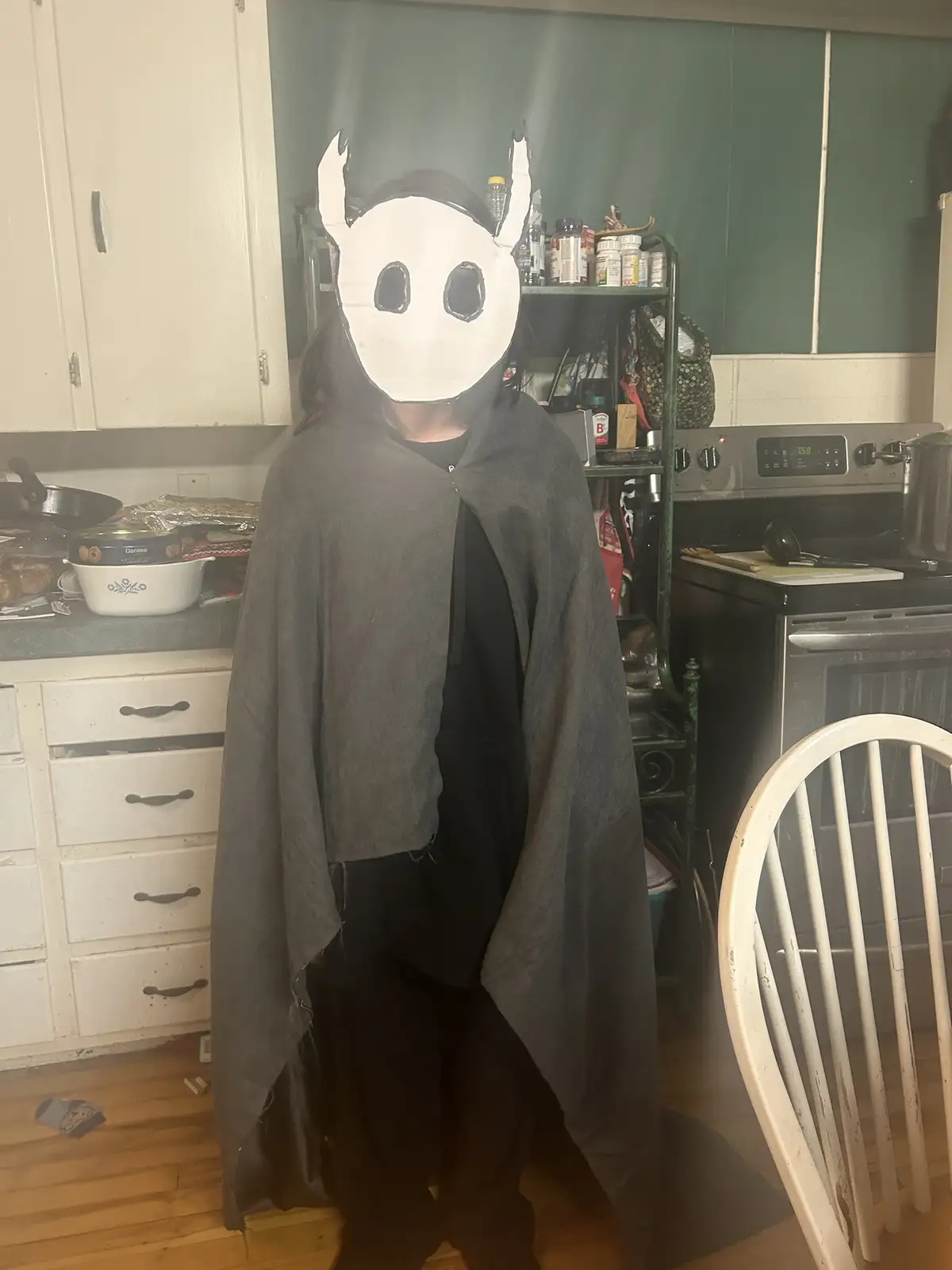 A person stands in a kitchen wearing a handmade white mask with black eyes and horns, draped in a dark grey cloak over black…