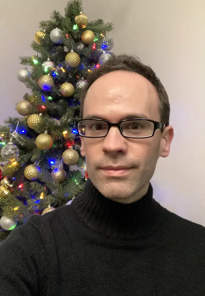 A man with glasses and a black turtleneck sweater stands in front of a decorated christmas tree with colorful lights and…