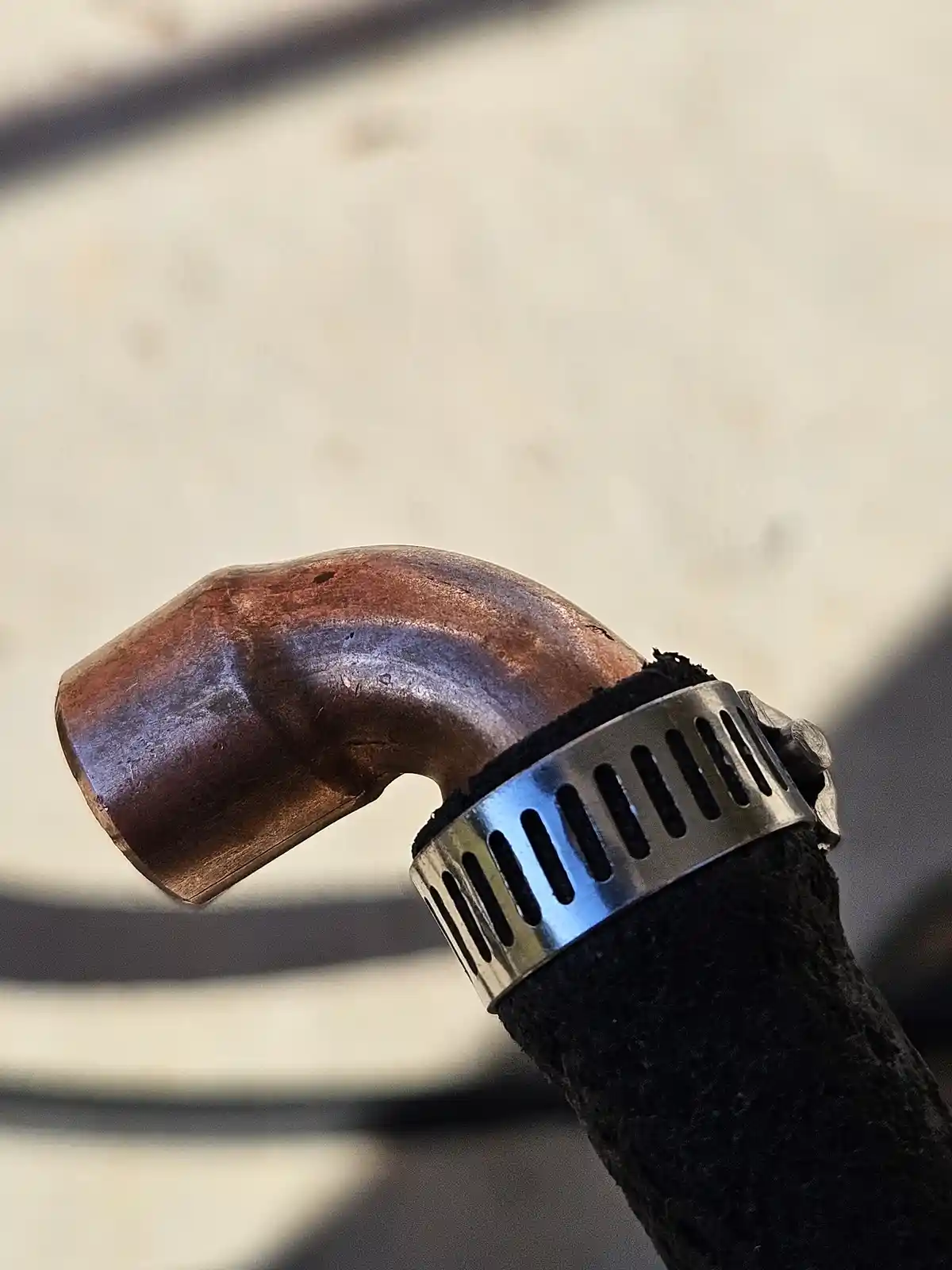 A close-up photo shows a copper pipe elbow joint connected to a black rubber hose with a metal clamp. The copper pipe has a…