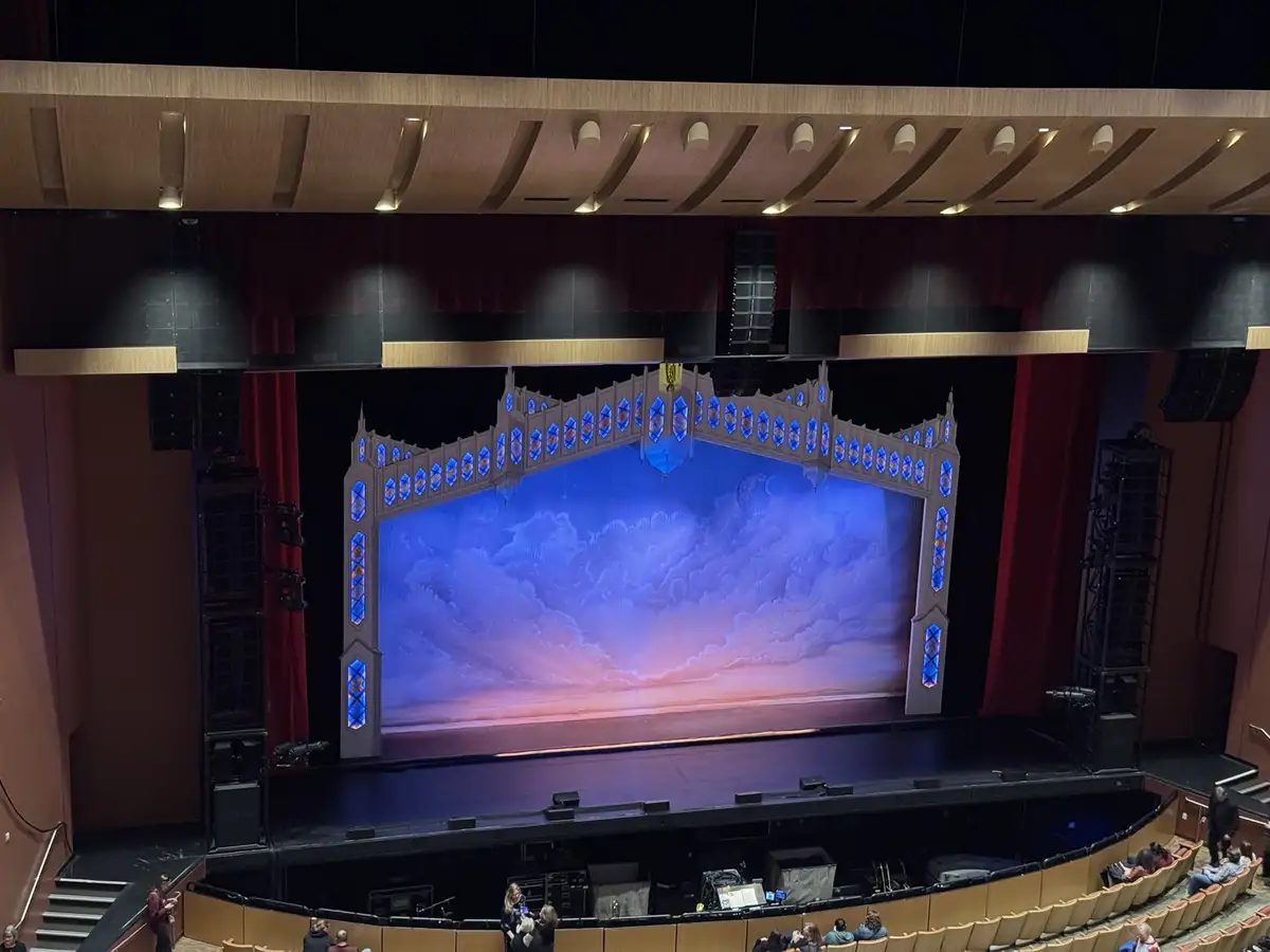 An empty theater stage with a large, ornate, blue-lit backdrop depicting a cloudy sky, surrounded by red curtains and stage…