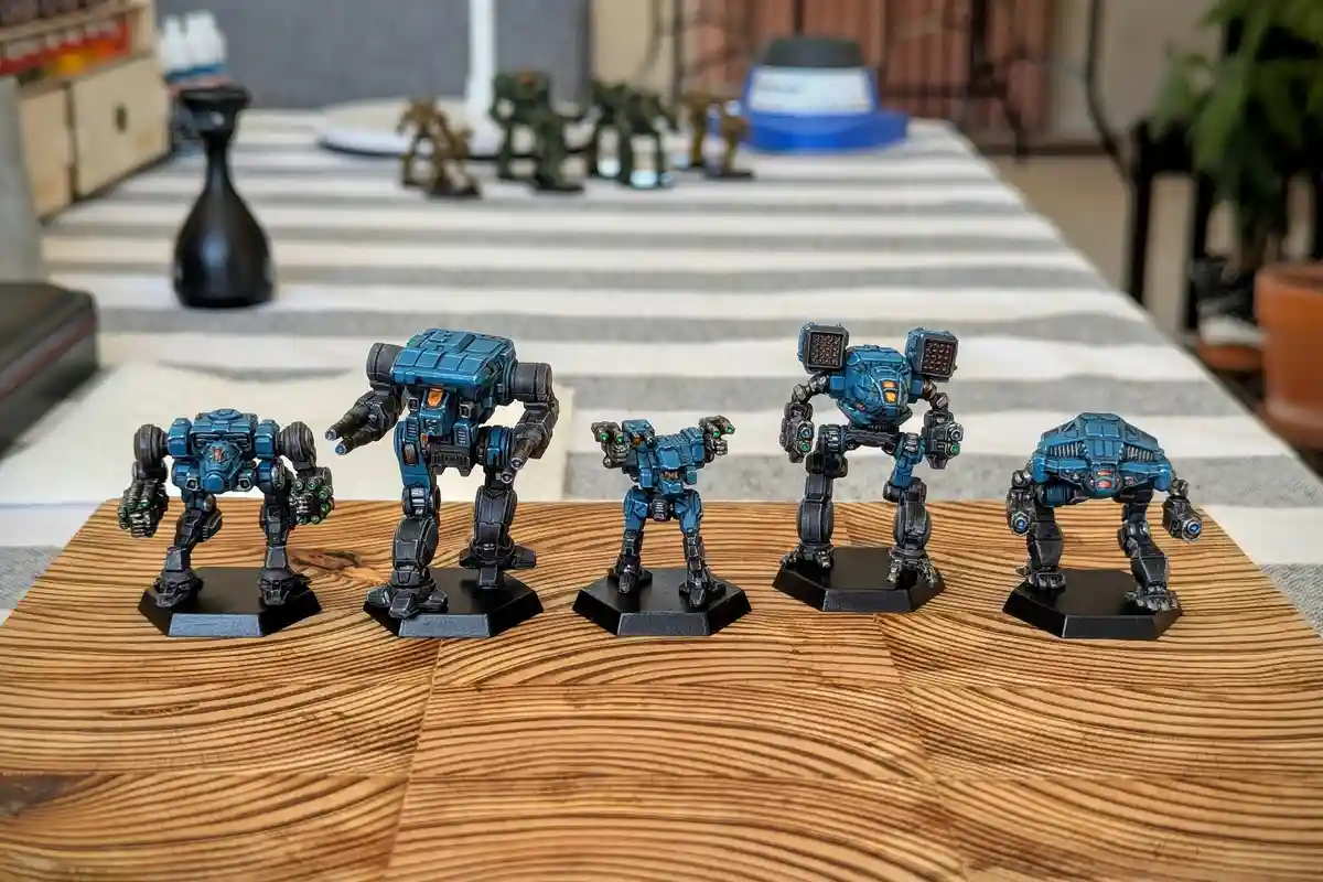 Five blue and black painted miniature war machines, resembling Titans from Warhammer 40k, are arranged in a row on a wooden…