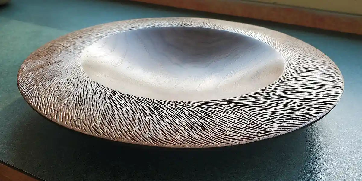 A close-up photo of a shallow, round, metallic-looking bowl with intricate, radiating engraved lines around its rim, resting…