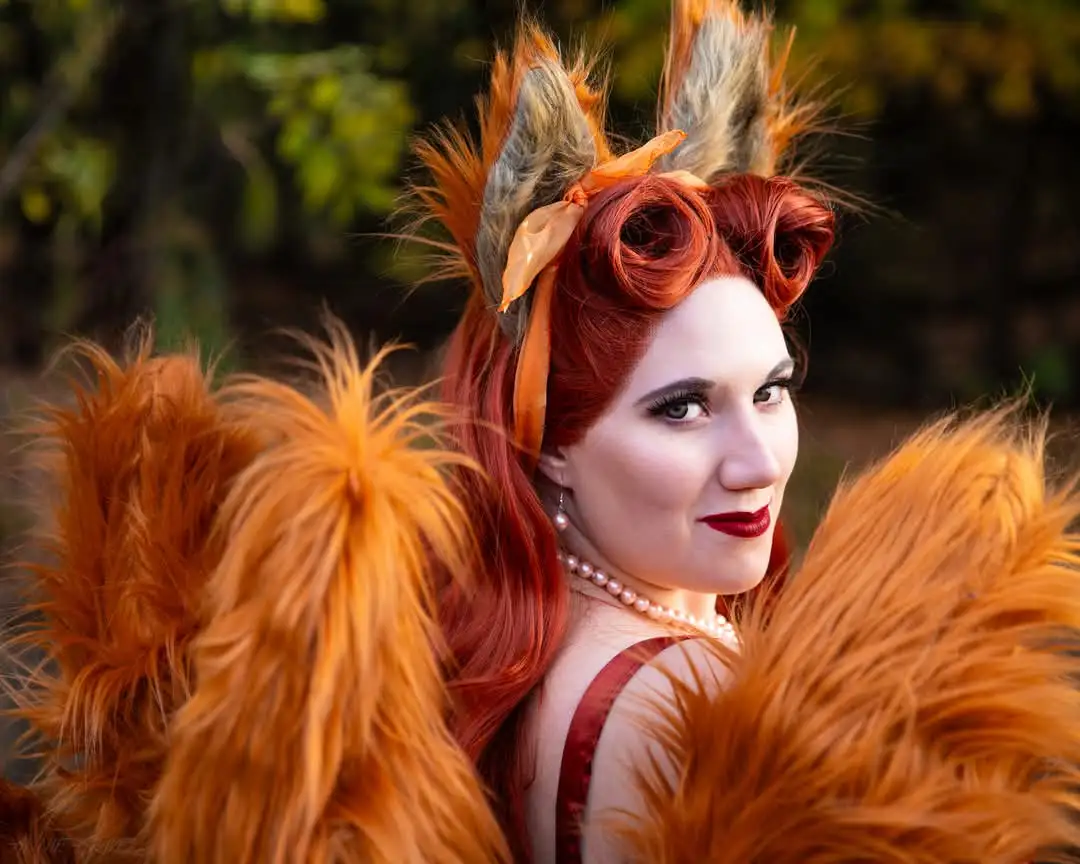 A woman with red hair styled in curls, wearing fox ears and a fluffy orange tail costume, looks over her shoulder with a sly…