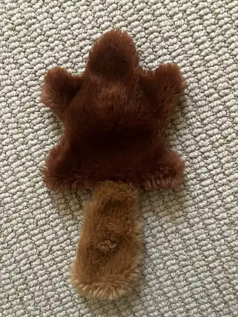 A brown, fluffy, paw-shaped dog toy with a handle lies on a textured white carpet. The overlaid text reads 'i'm not a fan of…