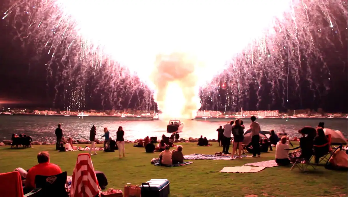 A crowd of people on a grassy waterfront watches a massive firework display, but a boat in the water is engulfed in flames…