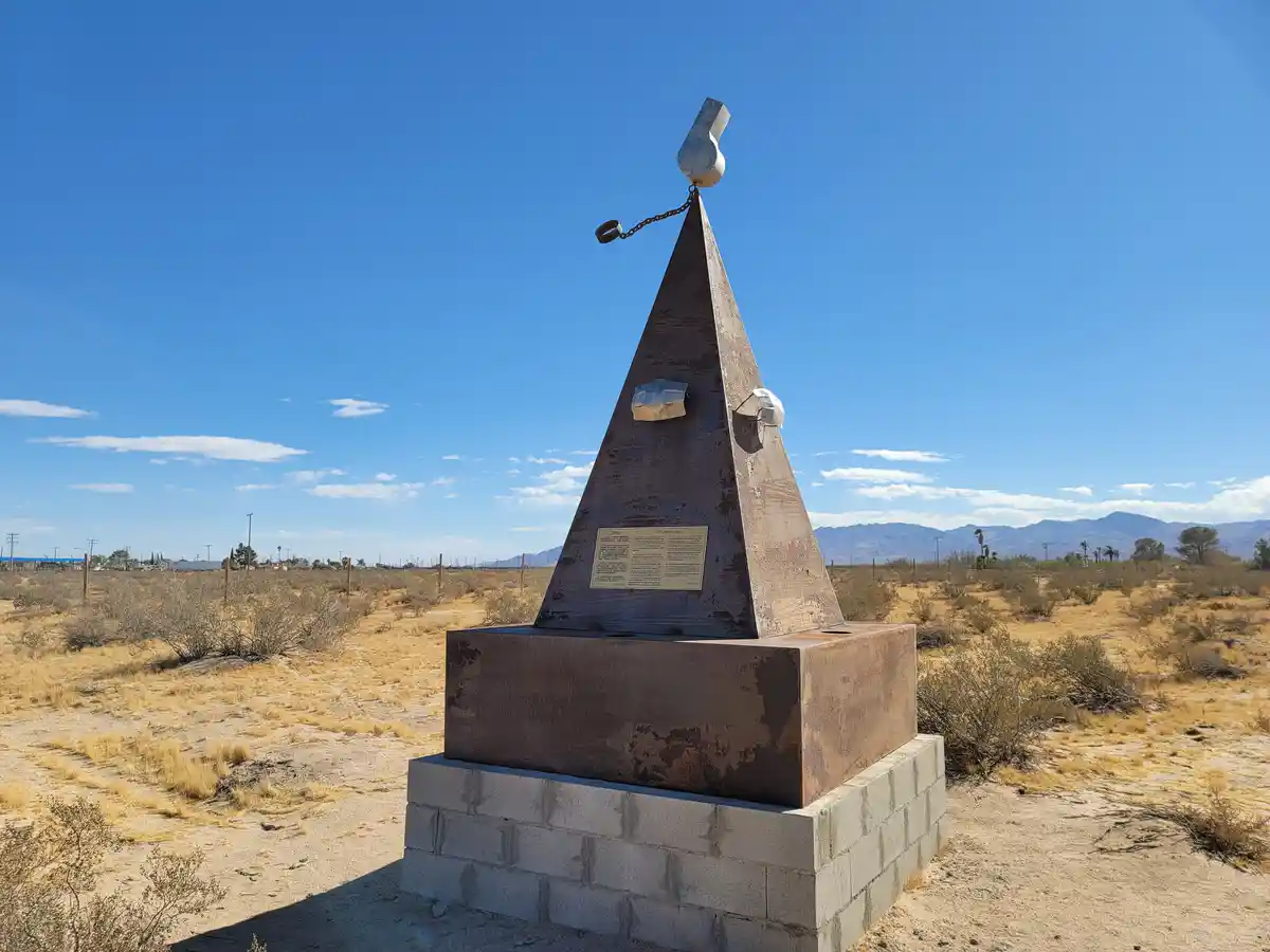 A rusted metal pyramid monument stands in a desert landscape under a clear blue sky, topped with a metal whistle attached by…