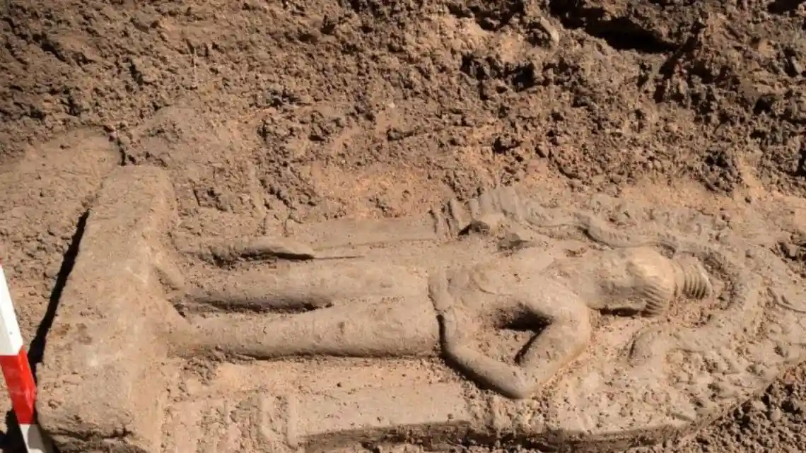 a weathered stone statue of a woman lying on her back is partially buried in dirt, with a red and white measuring stick…