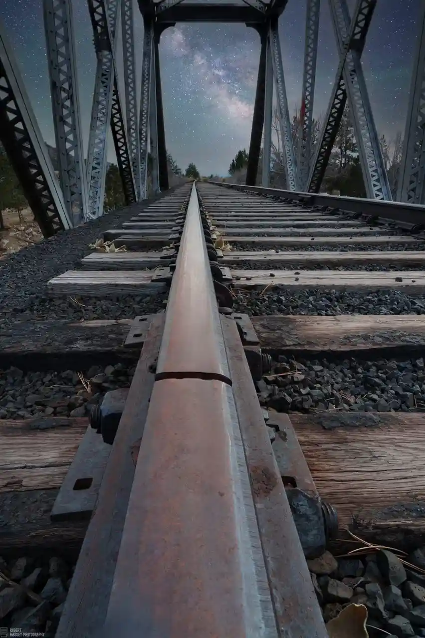 A low-angle shot looking down a rusty, weathered railroad track on a metal truss bridge at night, with the Milky Way galaxy…