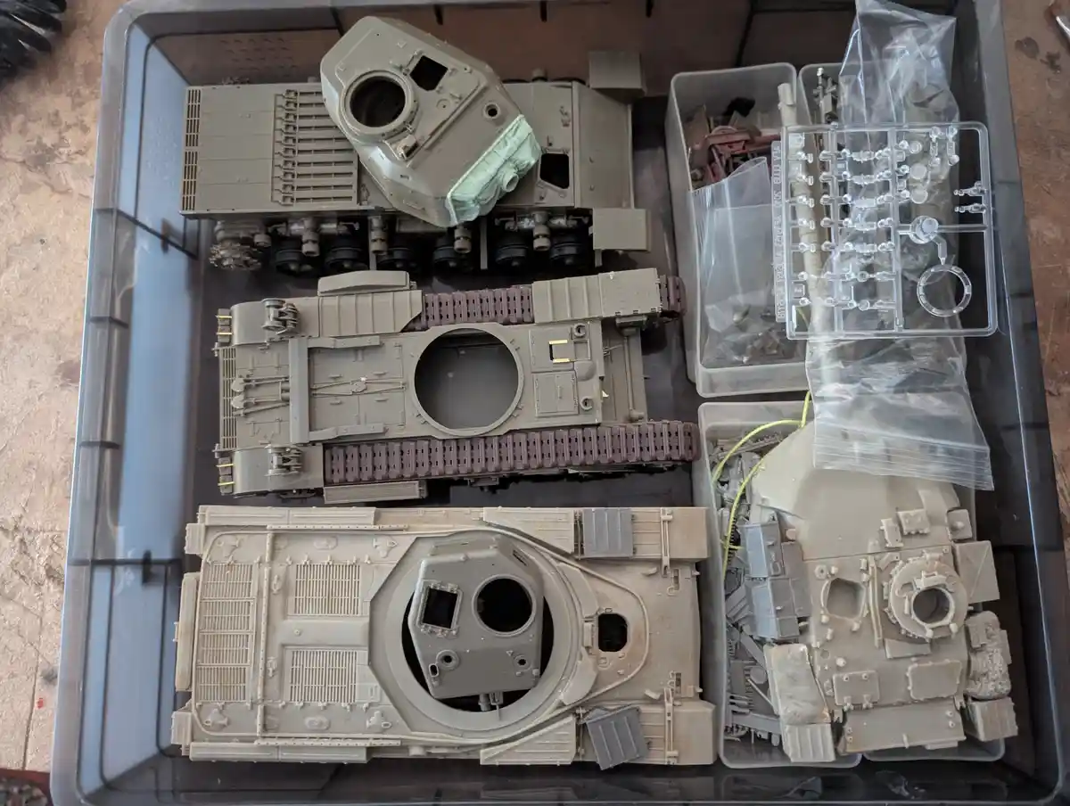 An overhead photo shows a plastic storage bin filled with unassembled model tank parts, including three large tank hulls in…