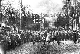 A black-and-white historical photograph shows a large crowd of people lining a cobblestone street, watching a man on…