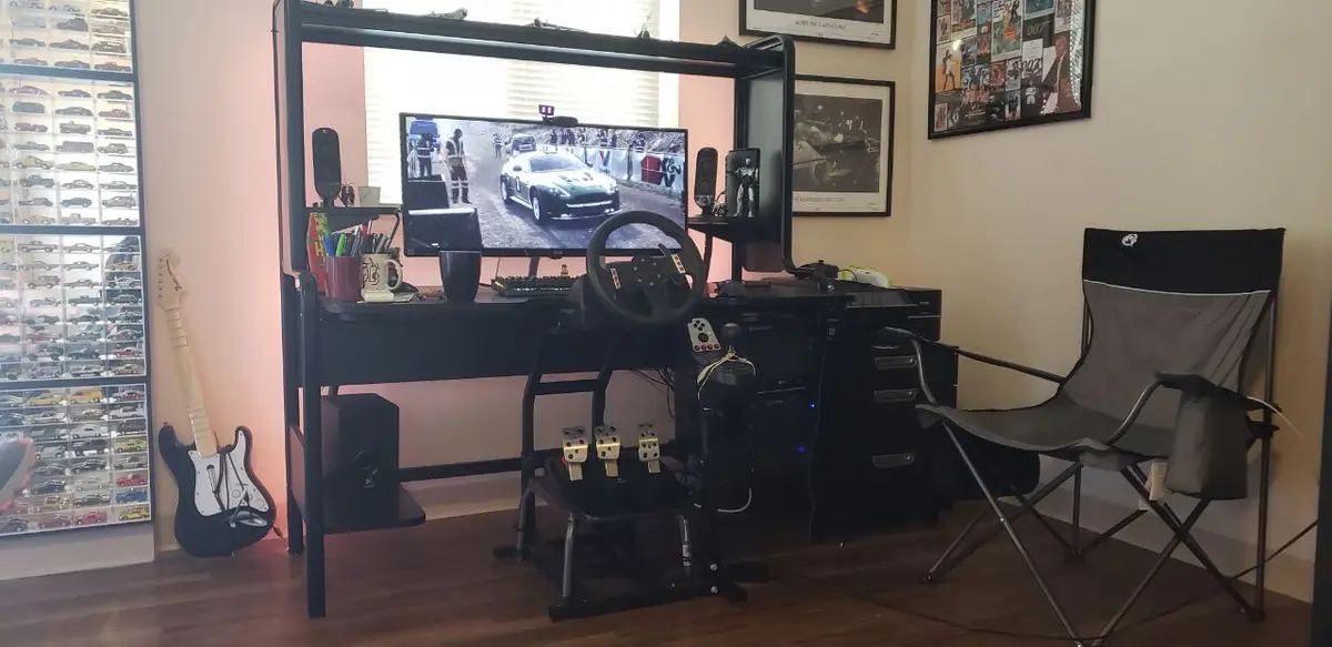 A room with a full racing simulator setup including a steering wheel, pedals, and large monitor showing a racing game. Next…