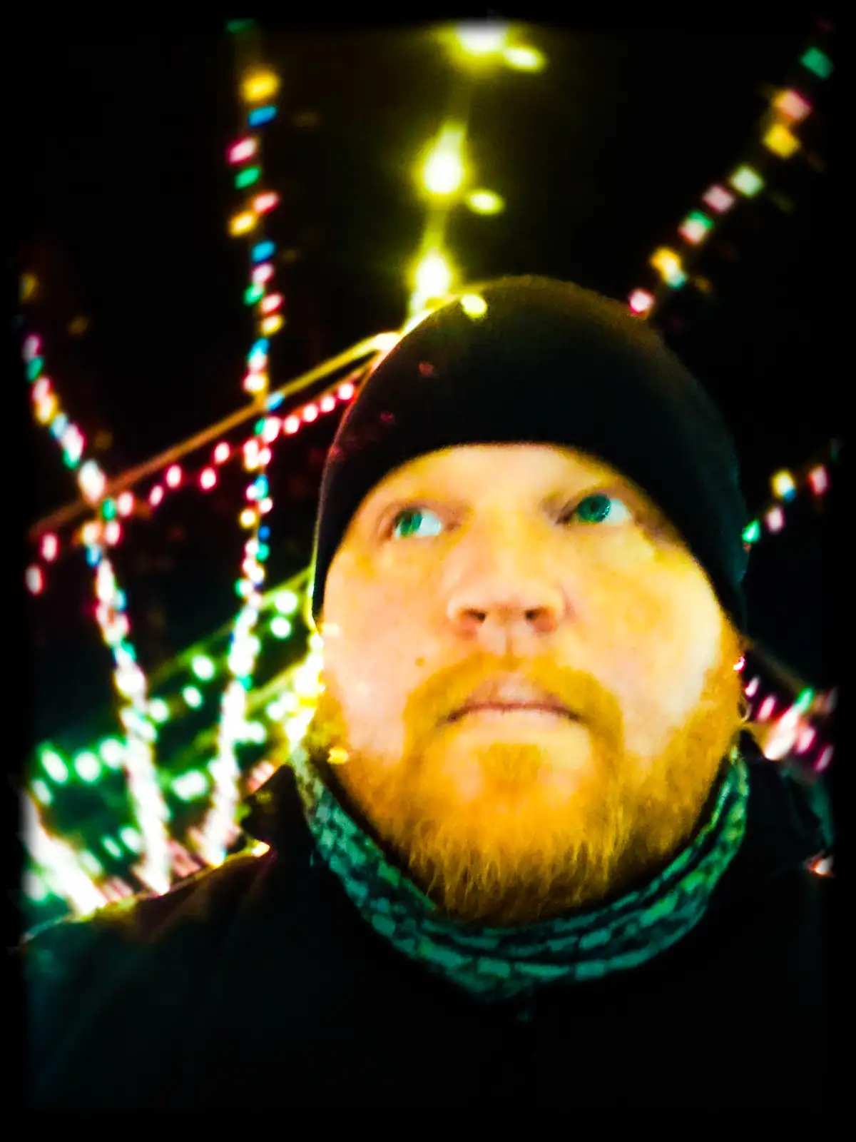 A man with a reddish beard and black beanie looks upward with a neutral expression against a dark background adorned with…