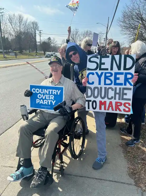 A man in a wheelchair holds a sign reading 'COUNTRY OVER PARTY' while smiling, next to another man holding a large sign that…