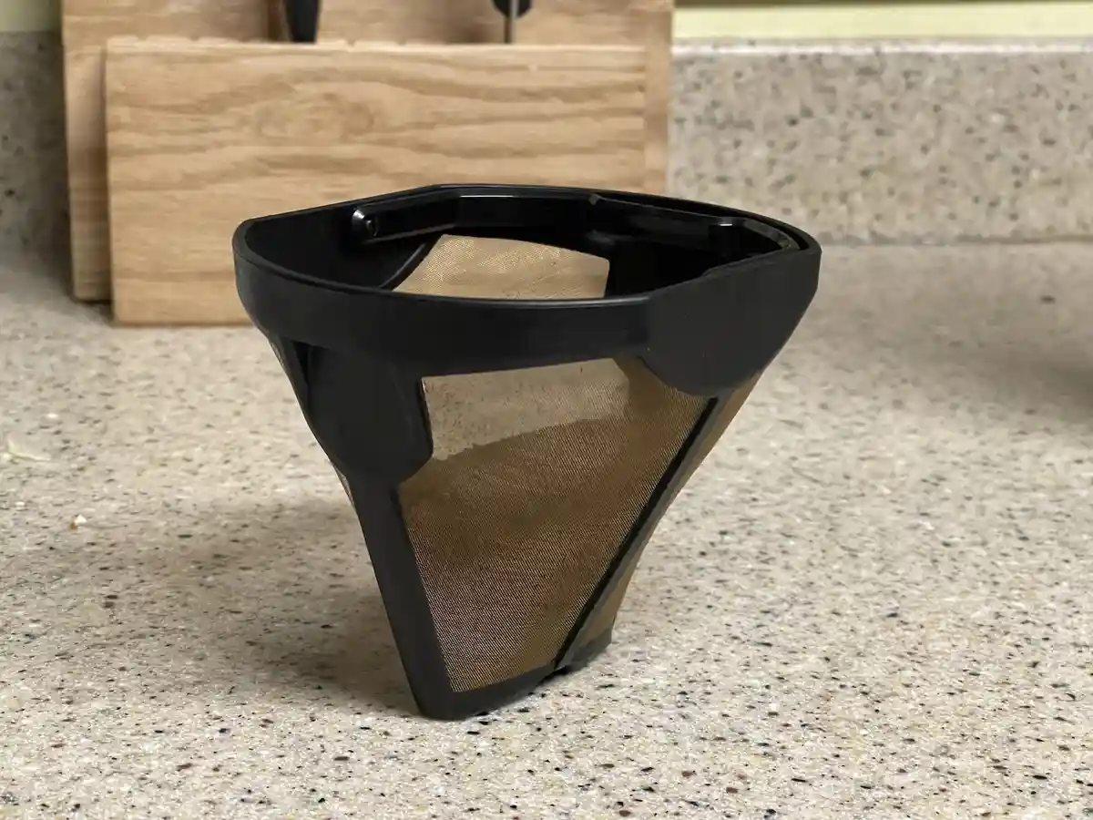 A black plastic coffee filter with a gold mesh interior sits on a speckled countertop, with a wooden knife block in the…
